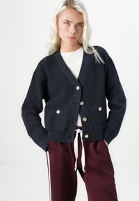 Navy ribbed cardigan with a V-neck, gold-tone buttons, and front pockets. Worn over a white top and paired with maroon pants.
