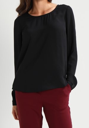 Vrouw die een effen zwarte blouse met lange mouwen draagt, gecombineerd met donkerrode broek, staand met één hand in de zak tegen een lichte achtergrond.