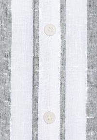 Camicia di lino con strisce verticali grigie e bianche, caratterizzata da un bottone beige al centro. Il tessuto presenta un aspetto strutturato.