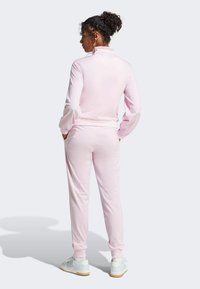 Rosa treningsdress med høy krage og ribbestrikkede mansjetter, med glatt tekstur og avslappet passform, kombinert med lyse joggesko.