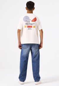 Camiseta de algodón blanca con formas geométricas coloridas y el texto "Confía en tu instinto" en la parte de atrás. Combinada con jeans de mezclilla azules.