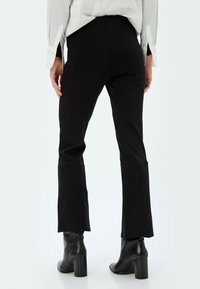 Mustat leveälahkeiset housut joustavasta kankaasta, joissa on korkea vyötärö, raaka helma ja sileä pinta, yhdistettyinä näyttäviin mustiin korkokenkiin.