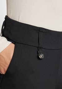 I pantaloni neri a vita alta presentano un tessuto liscio e elasticizzato, una chiusura con bottone e tasche laterali, con una silhouette affusolata e cuciture minime.