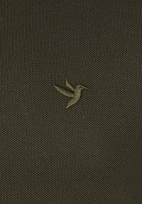 Olivengrüner, strukturierter Stoff mit einem kleinen, gestickten Kolibri-Logo in einem etwas dunkleren Grünton.