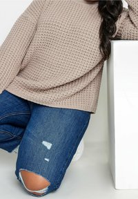 Kvinna på knä iförd trasiga blå jeans, beige stickad tröja och vita sneakers, lutad med armen på en vit block mot en enfärgad bakgrund.