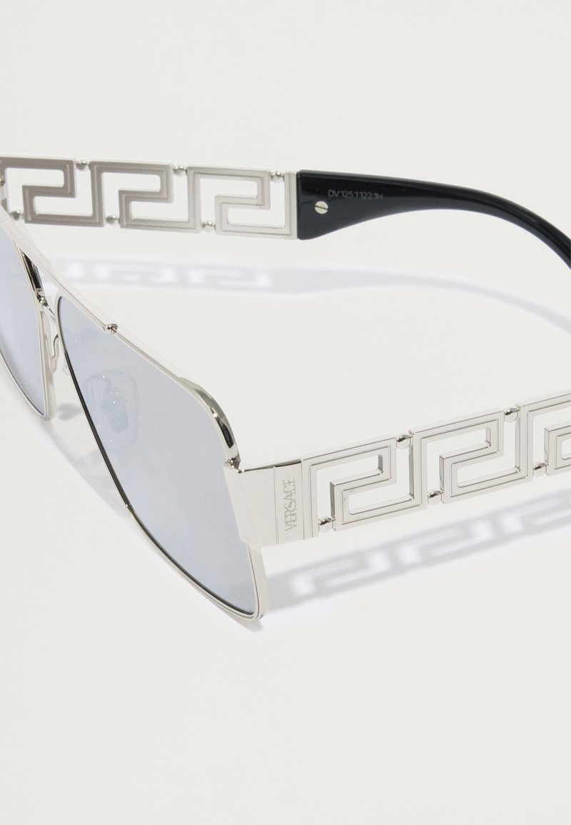 Sonnenbrille mit rahmen in silberner Optik, geometrischem Muster an den Bügeln und grauen Gläsern. Schwarze Bügel mit Branding auf der Innenseite. Rechteckige Form.