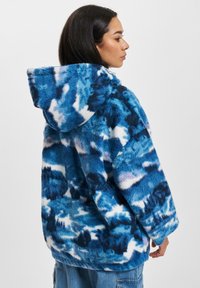 Faux fur hettejakke i blåtoner; har abstrakte sky-lignende mønstre med teksturer og en avslappet silhuett.