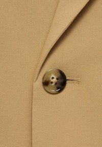 Tessuto beige dalla superficie liscia, con un grande bottone rotondo marrone a quattro fori, attaccato a un capo di abbigliamento sartoriale.