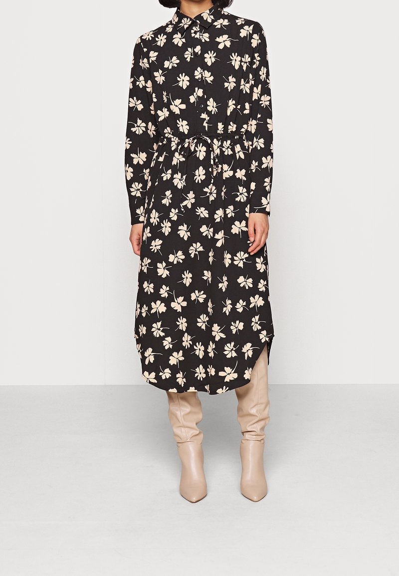Femme portant une robe midi noire à manches longues avec imprimé floral beige et des bottes beige montantes, debout devant un fond uni.