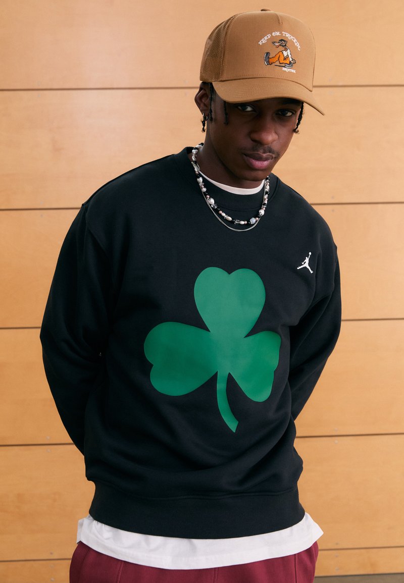 Μαύρο φούτερ με μεγάλο πράσινο σχέδιο τριφυλλιού, συμπληρωμένο με μικρό λευκό λογότυπο στο στήθος. Φορεμένο με μπεζ καπέλο και πουκάμισο σε στρώσεις.