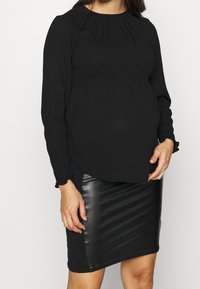 Černý top s dlouhým rukávem a nařasenými sklady, zakončený elastickými manžetami, v kombinaci s přiléhavou černou sukni z faux kůže ve stylu tužky s diagonálním žebrováním.
