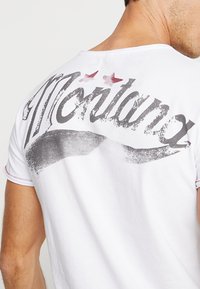 Camiseta de algodón blanca con un estampado gráfico desvanecido que presenta "Montana" en negro y estrellas rojas decorativas en la parte espalda. Mangas cortas y cuello redondo.
