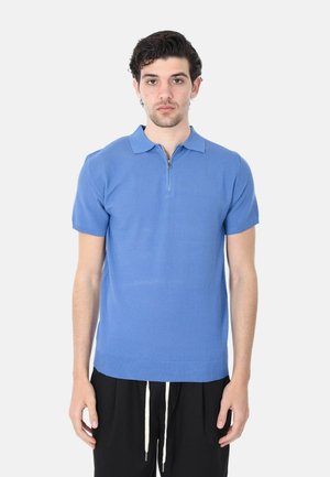 Giovane uomo in piedi, indossa una polo a maniche corte di colore blu in maglia con una cerniera frontale e pantaloni neri con coulisse bianche, rivolto verso davanti.