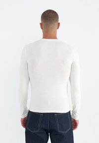 Hosszú ujjú, krémszínű felső, puha anyagból készült, slim fit szabású, kerek nyakkivágással, amelyet sötétkék farmernadrággal párosítottak, hátsó zsebekkel.