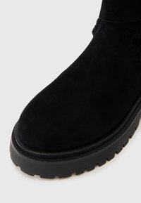 Botă din suede negru, cu vârf rotunjit și talpă groasă, având o căptușeală confortabilă vizibilă la marginea superioară.