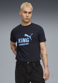 T-srajica iz navadne bombaža v mornarsko modri barvi, krasijo jo svetlo modra grafika "KING" in logotip Puma, okrogli izrez in kratki rokavi. Ustrezajo ji črne kavbojke.