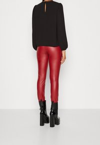 Personne portant un pantalon en cuir rouge taille haute, un chemisier noir à manches longues et des bottines plateformes noires, debout devant un fond uni.