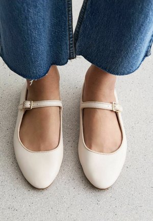 Zapatos Mary Jane de cuero beige claro con punta redondeada, pequeño tirante y detalle de hebilla, combinados con pantalones de mezclilla anchos.