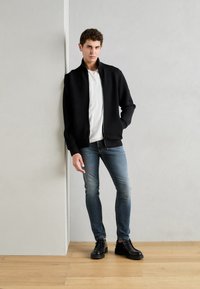Fekete cipzáras dzseki, fehér ing, világoskék slim fit farmernadrág és fekete cipő. Simább anyag, laza illeszkedés, egy világos falnak támaszkodva.