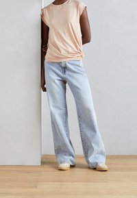 Top corto de melocotón claro con detalle de nudo torcido, combinado con jeans de pierna ancha azul claro y zapatos beige sobre un suelo de madera.