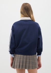 Felpa blu navy con colletto bianco a contrasto, dotata di polsini a costine e strisce laterali, abbinata a una gonna pieghettata a quadri marrone.