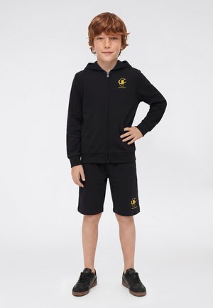 Ragazzo con capelli rossi in piedi con una mano sul fianco, indossa una felpa nera con zip e pantaloncini abbinati con loghi gialli e scarpe da ginnastica nere.