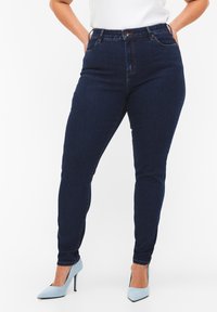 Jeans a vita alta di colore blu scuro con una vestibilità slim, design a cinque tasche, cuciture a contrasto e chiusura con bottone. Abbinati a tacchi di colore blu chiaro.