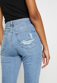 Jeans de mezclilla de un azul claro con cintura alta, silueta ajustada y detalles desgastados en los bolsillos traseros. Textura suave, costura estándar.