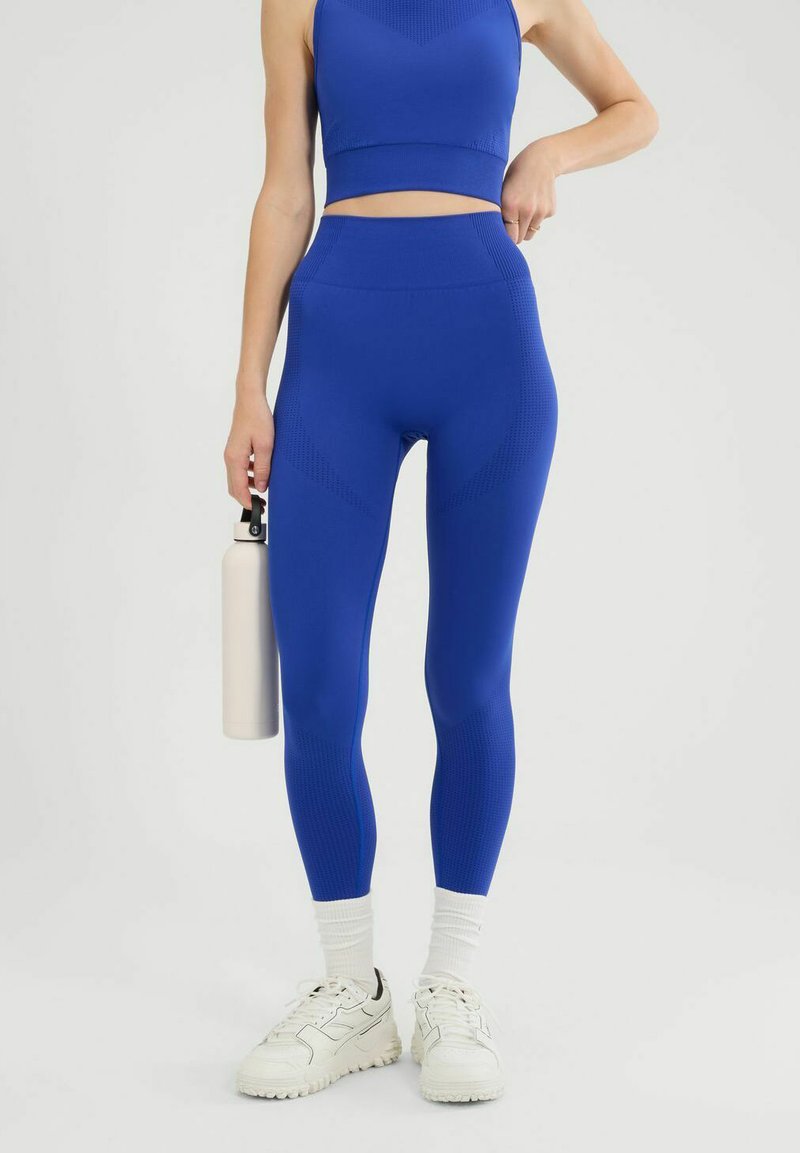 Wysokie niebieskie legginsy z teksturowanymi panelami i dopasowanym topem. Biała butelka wody trzymana w ręce oraz białe sneakersy dopełniają całość.