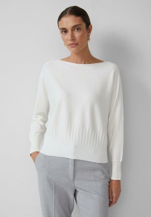 White knitted sweater with a boat neckline, ribbed hem, and long sleeves. Paired with light gray trousers, creating a simple, sleek look.