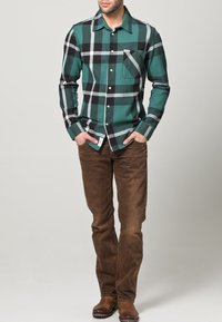 Camisa de franela a cuadros verdes y negros con cierre de botones y bolsillo en el pecho, combinada con jeans marrones y botas marrones.