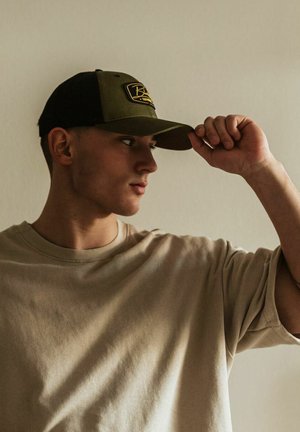 Olive green and black cap with a logo patch, worn at an angle. Light beige, short-sleeved shirt with a soft texture, worn casually.