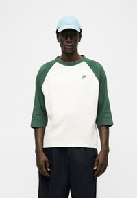 Άνδρας που φοράει λευκό και πράσινο πουκάμισο τριών τετάρτων μανίκι της Nike, μαύρο παντελόνι και ανοιχτό μπλε καπέλο Nike, στέκεται μπροστά σε λευκό φόντο.