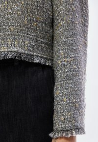 Γκρι υφασμάτινο σακάκι με υφή tweed, με ξεφτισμένα άκρα και χρυσές κηλίδες, σε συνδυασμό με σκούρο τζιν παντελόνι. Κοντινό πλάνο λεπτομέρειας στο μανίκι.