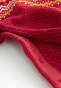 Tejido de punto rojo con un patrón texturizado, con botones visibles y una textura suave y lisa. Acentos de amarillo y blanco en el patrón.