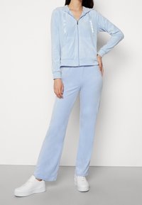 Ljusblå velour träningsdräkt med en zip-up huvtröja och matchande vida byxor. Har vita snoddar och en mjuk, slät textur.