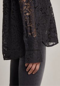 Camicia nera in pizzo con motivo floreale, maniche lunghe e polsini con bottoni. Il tessuto è semitrasparente con una finitura testurizzata.