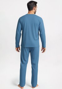 Man staat blootsvoets met zijn rug naar de camera, gekleed in een bijpassende blauwe lange mouwen top en broek voor een eenvoudige witte achtergrond.