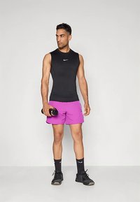 Hombre atlético con camiseta sin mangas negra, pantalones cortos rosas, calcetines y zapatillas negras de Nike, sosteniendo una botella de agua, de pie contra un fondo simple.