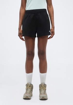 Osoba nosiaca čierne športové šortky, biele členkové ponožky a béžové turistické topánky, stojaca pred jednoduchým bielym pozadím.