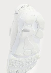 White sports shoe with textured fabric upper, thick foam sole, unique tread pattern, and a Velcro strap for secure fit.