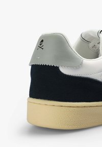 Sneaker mit einer oberen Hälfte aus weißem Leder, einem navyblauen Wildlederpaneel, einer grauen Lederfersenlasche mit einem Totenschädel- und Knochen-Logo sowie einer cremefarbenen Gummisohle.