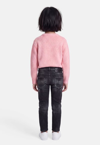 Παιδί με κοντά σκούρα μαλλιά στέκεται πλάτη, φορώντας ροζ πουλόβερ, μαύρα ξεβαμμένα τζιν και λευκά αθλητικά παπούτσια σε ανοιχτό φόντο.