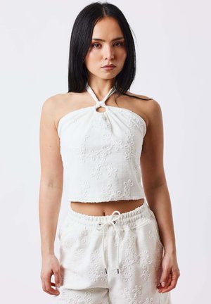 Joven con cabello negro lacio que lleva un top corto halter blanco con textura floral y pantalones a juego con cordón ajustable.