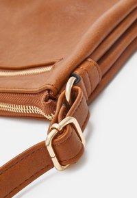 Detailní záběr rohu hnědé kožené tašky se zlatou kovovou sponou a zipem na bílém pozadí.