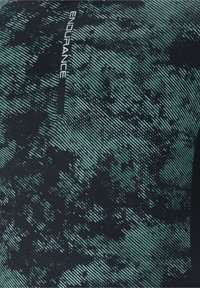 Gros plan sur un tissu à motifs verts et noirs avec une fermeture éclair noire verticale portant l’étiquette "ENDURANCE" en texte blanc.