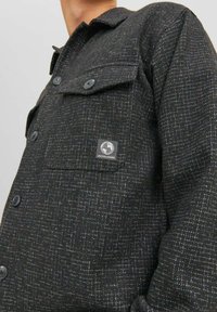 Giacca camicia nera con motivo, realizzata in un tessuto testurizzato, caratterizzata da dettagli in filo blu e bianco, una tasca sul petto e bottoni neri. Logo del marchio visibile.