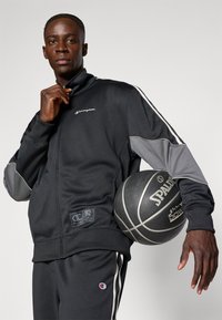 Μαύρη αθλητική φόρμα με γκρι λεπτομέρειες και λογότυπο Champion, που περιλαμβάνει μπουφάν με φερμουάρ και παντελόνι με ρίγες; κρατώντας μια μπάλα μπάσκετ Spalding.