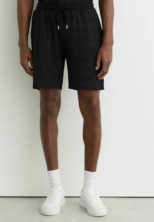 Mężczyzna w czarnych, teksturowanych szortach, białych skarpetkach do kostek i białych trampkach, stojący na tle jasnej ściany.