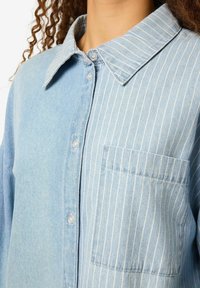 Chemise en jean bleu clair avec un devant à boutons, un col classique et une poche sur la poitrine gauche arborant des rayures verticales blanches. Texture douce.